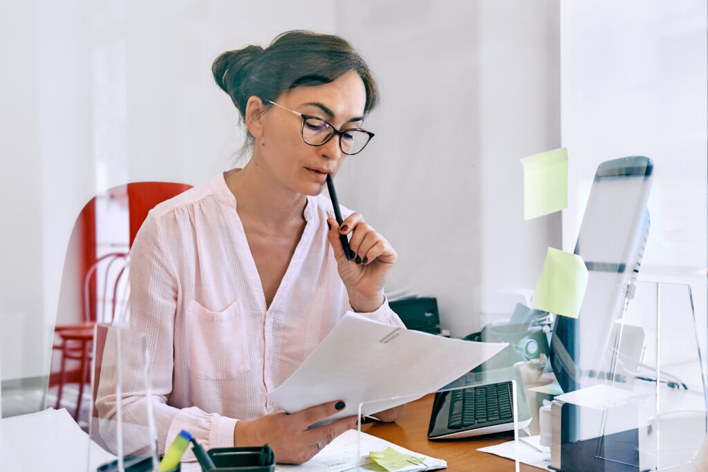 Attentive female bookkeeper check accounts bills. Middle-aged woman social worker counting grant subsidy rates, reviewing the company's financial data at office workplace with plexiglass separator.